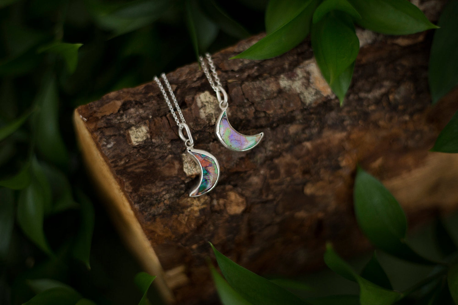 Two sterling silver and Monarch opal crescent moon necklaces laying side by side, back to back in a woodlands scene